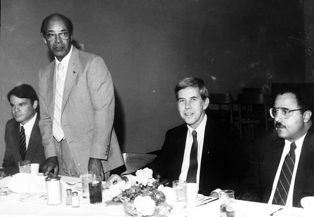 W. T. Ray stands at a table. Three other men are seated next to him.