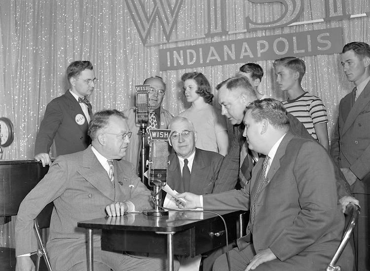A group of four men huddle around a microphone. Two men and four teenagers stand in the background next to another microphone.