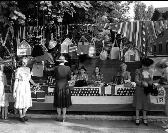 Women are seated at a table selling cloth purses and clothespin bags. The table is decorated with patriotic fabric.