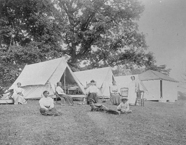 Several women and some children are resing on the lawn in front of raised camp tents.