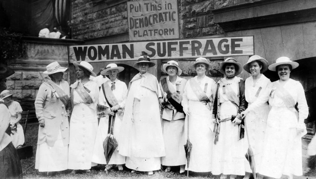 A group of women stand in front of a sign that reads "Woman Suffrage."