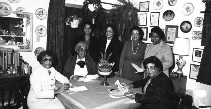 A group of Black women sit and stand around a dining room table. 