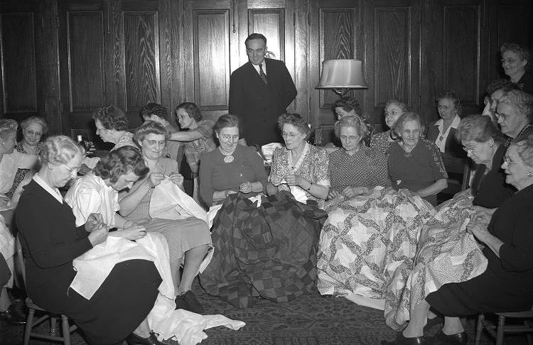 A group of women are sitting in a room and hand sewing quilts. 