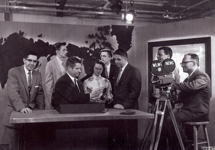 A group of people are gathered on a news set. A world map decorated the background. A camera is pointed on the set. 