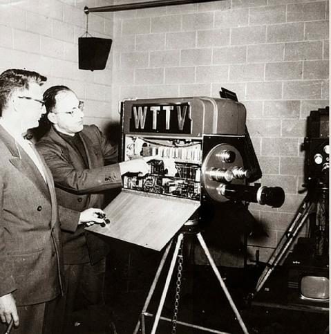 Two men look at a television camera with the side panel open and the inner workings exposed. One of the men points to a part inside the camera.