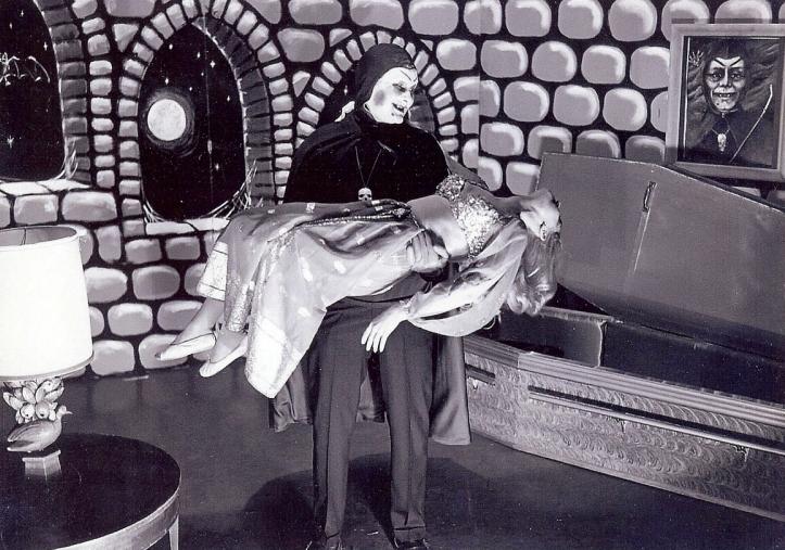 A man dressed in ghoulish costume holds a woman. The background is decorated to look like a castle and a coffin is sitting on the floor. 