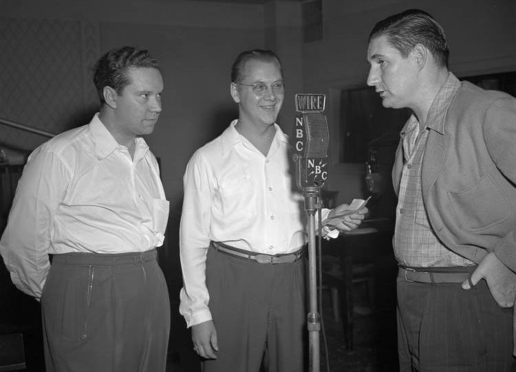 Three men stand around a microphone. 