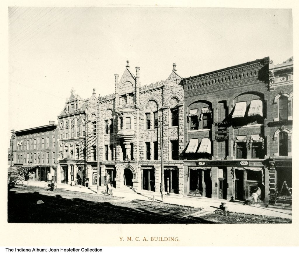 The YMCA building is located in the middle of a block of buildings. Signs can be seen for Wilson's, C. F. Schmidt's Wiener Beer, Henry Smith Sample Room Restaurant, Norb. Landgraf, and June's Famous Oyster & Chop House.