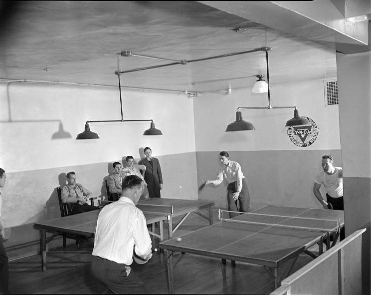 Two sets of men play table tennis. Four men watch from the edge of the room. 