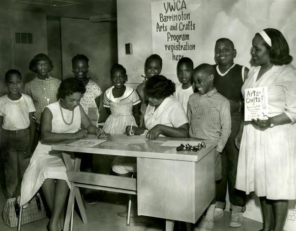 	 Boys and girls are shown registering for the YWCA Arts and Crafts Program. One of the women holds a sign saying it starts June 28.