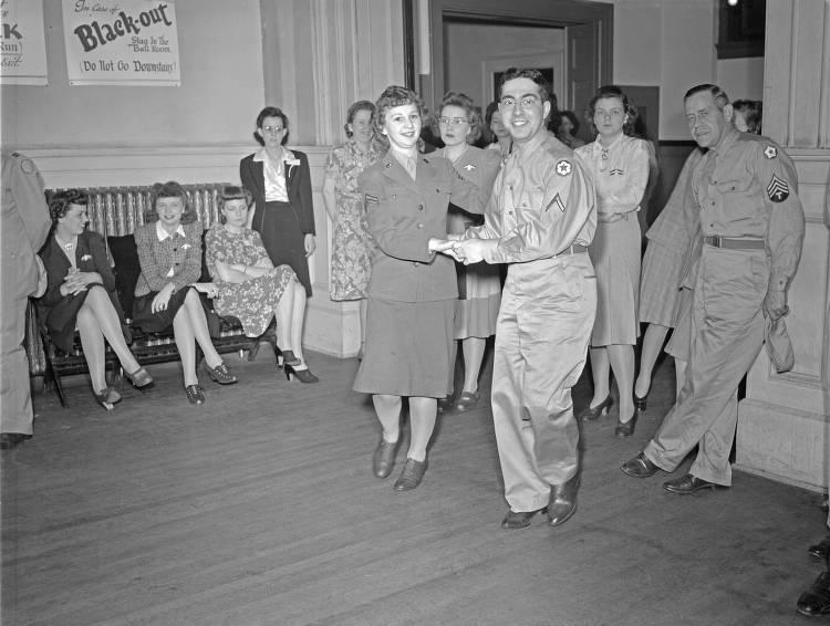 A uniformed couple dances in the middle of a room. Several women sit and stand along the edges of the room. A man is leaning against a wall.