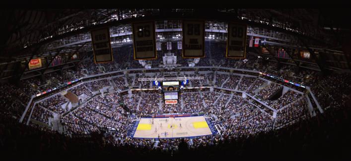 View of the interior of a large arena from the upper seats showing a basketball court (game in progress) encircled by tiered rows of sets full of spectators.