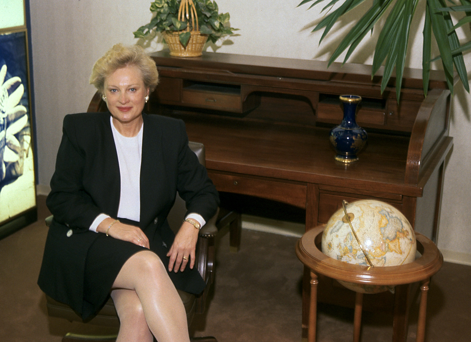 A woman in a business suit sits next to a desk.