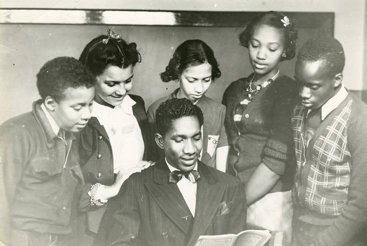 Five students stand behind a man who is seated and reading from a book.