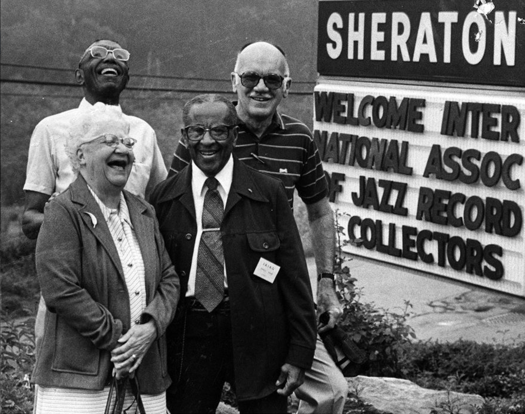 Four people stand next to a sign outside that is welcoming a jazz association.