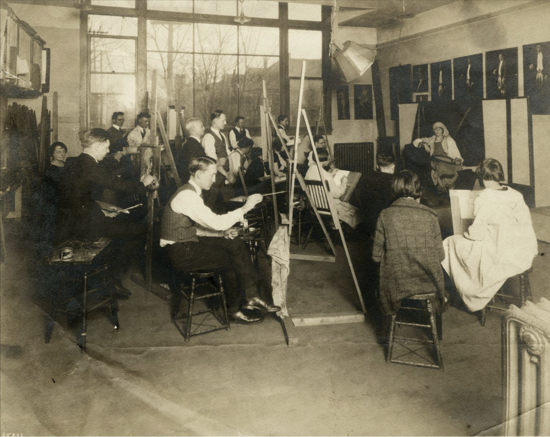Several students sit at easels and paint in a large classroom. Their model poses at the front of the classroom.