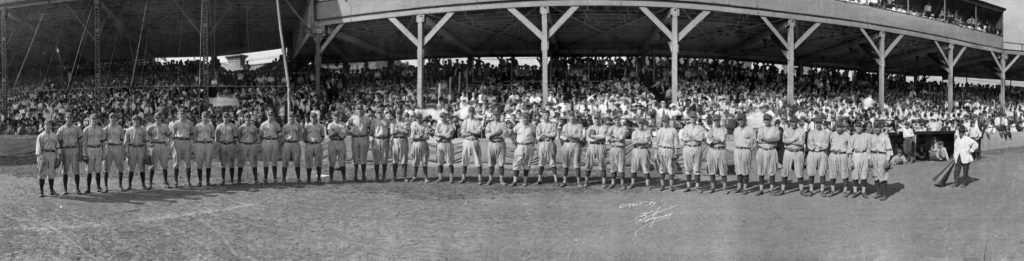 A long row of baseball players are lined up in front of a covered grandstand full of people.