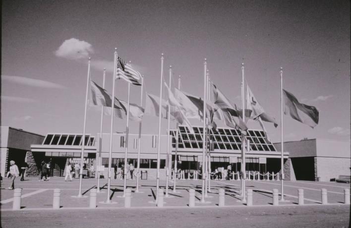 An entrance with a plaza filled with flags. People walk around the entrance.