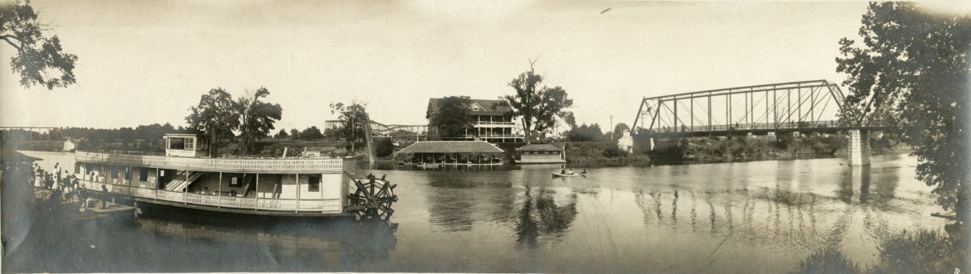 View of people boarding a riverboat across from Riverside Amusement Park. A roller coaster and a bridge over the White River are visible.