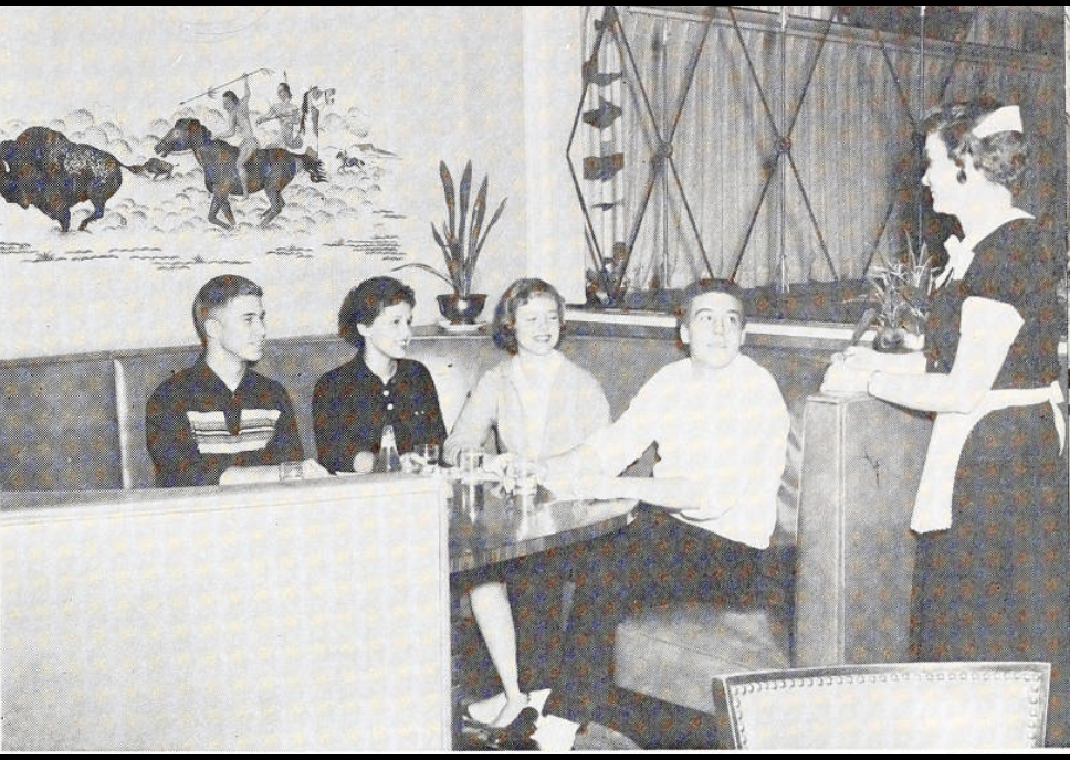A group of young adults sit in a circular booth. A waitress is talking to them.  
