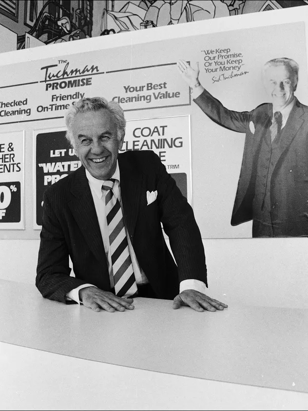 Sid Tuchman sits at a counter. An ad for Tuchman Cleaner's covers the wall behind him.