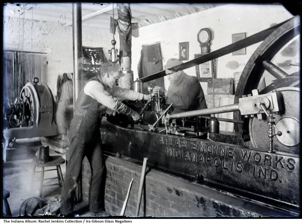 Men working on a stationary engine with a wheel and belt that is stamped "Atlas Engine Works, Indianapolis, Ind."