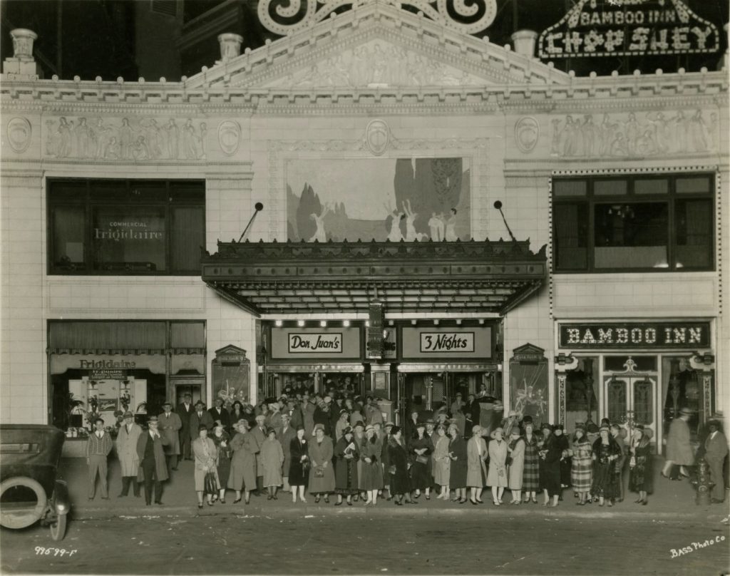 A large crowd of people stand on the sidewalk in front of a theater building. A restaurant, the Bamboo Inn, is part of the building. 