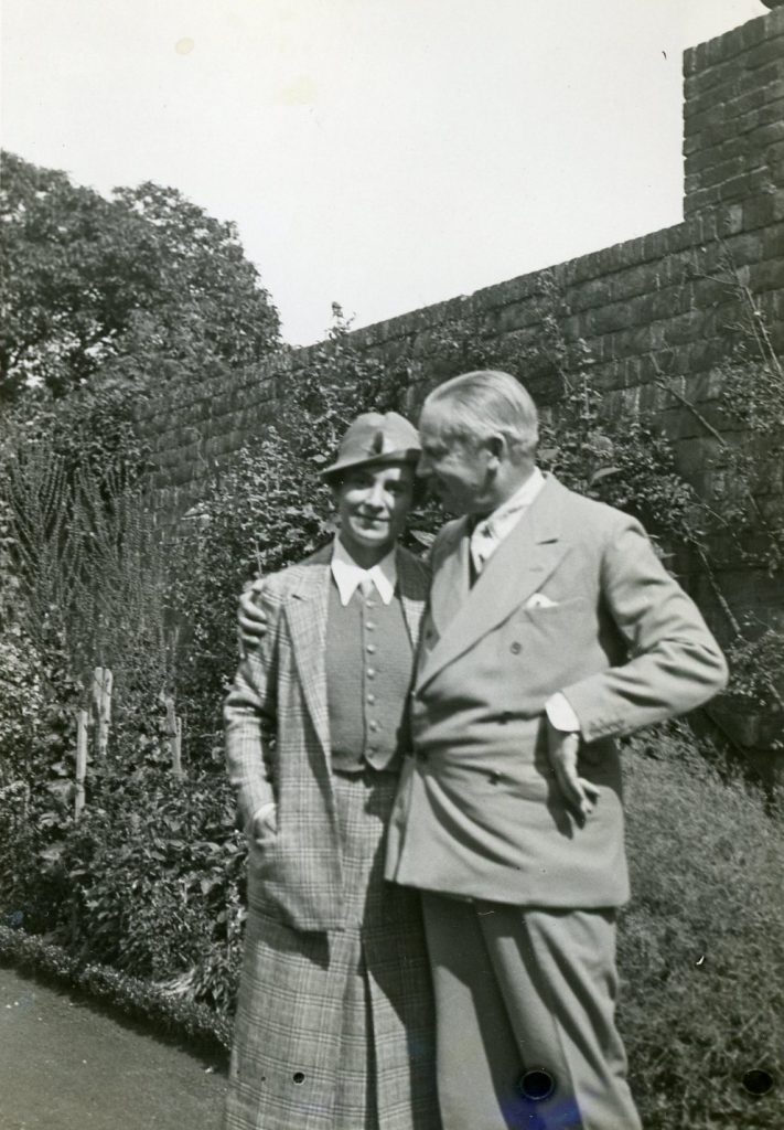 A man wraps his arm around the shoulders of a woman. They are standing outside, in front of a brick wall.