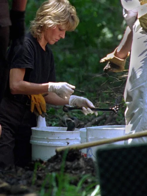 A woman wearing gloves examines a human bone. She is kneeling on the ground over multiple buckets that also contain human bones.