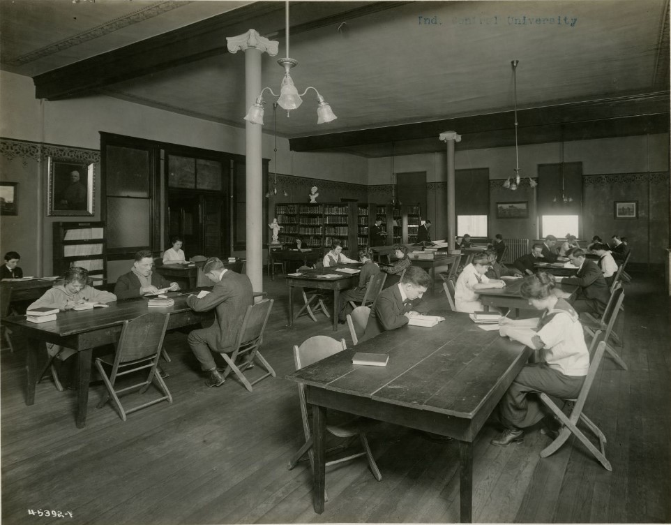 Students are working and reading at wooden tables in the library. The boys are wearing suits while the girls all wear skirts and blouses. Bookcases are in the back left corner of the room.