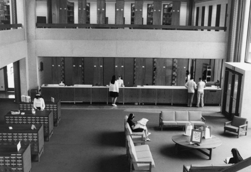 A wide view of the interior of a library. One person is going through the card catalogue, a nun is seated on a couch, and several individuals are getting help at the front desk. 