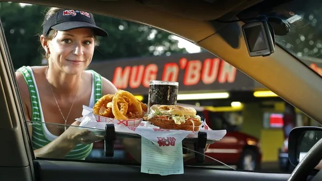 An employee wearing a Mug n Bun visor delivers a tray of food through the window of a customer's car. 