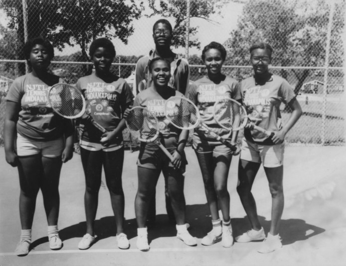 A group of young adults pose on a tennis court with their rackets. 