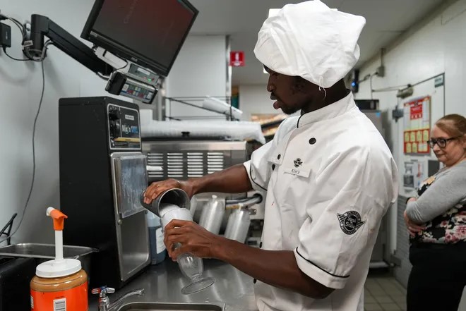 An employee dressed in a chef's uniform pours a vanilla shake in a glass.