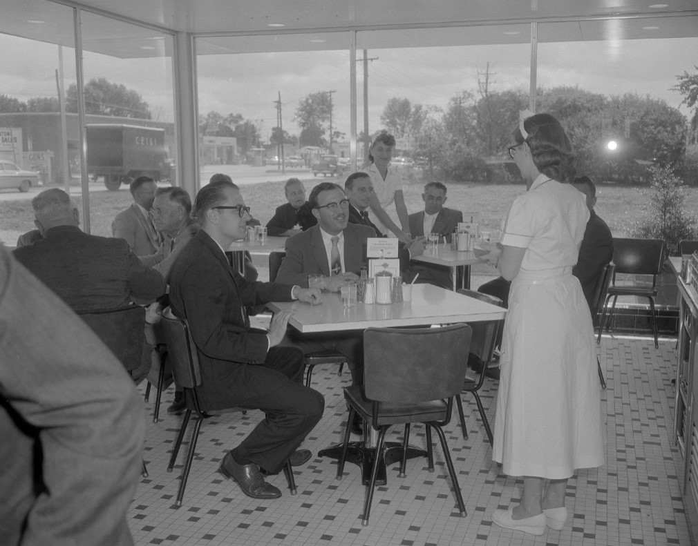 A waitress takes the order of two men seated at a table. Another waitress attends to a table of customers in the background. 