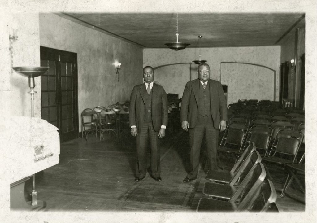 Two men dressed in suits stand in a funeral home. Chairs are set up in the room and there is a closed casket in the corner. 