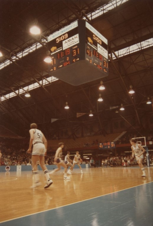 Players are on a basketball court in mid-game. A crowd of people watch from the lower levels of the stands. The upper levels of the stands are empty. 