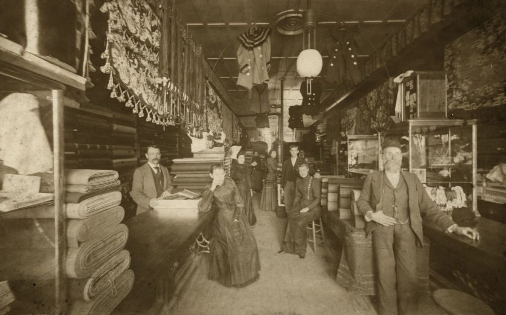 Several men and women turn to face the camera in a dry goods store.  The women wear close-fitting dresses with no bustle and slightly puffy sleeves, and the men wear three-piece suits with cutaway jackets.  Various textiles, ready-to-wear clothing, and sundries can be seen for sale.