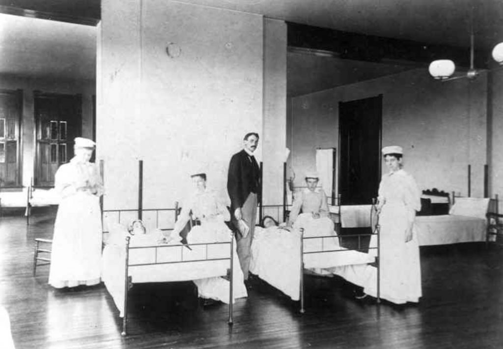 Four nurses stand next to two hospital beds with a patient in each bed. A man stands in the middle of the two beds. 