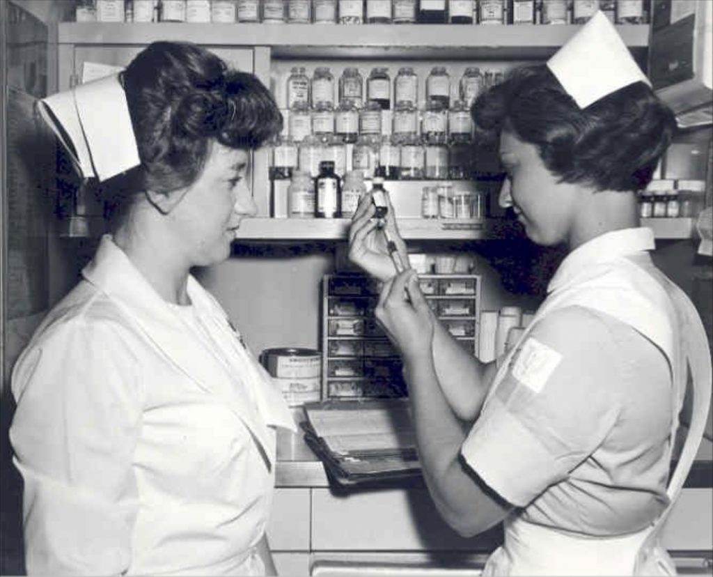 Two nurses stand in front of a medicine cabinet. One nurse is drawing fluid into a syringe. 