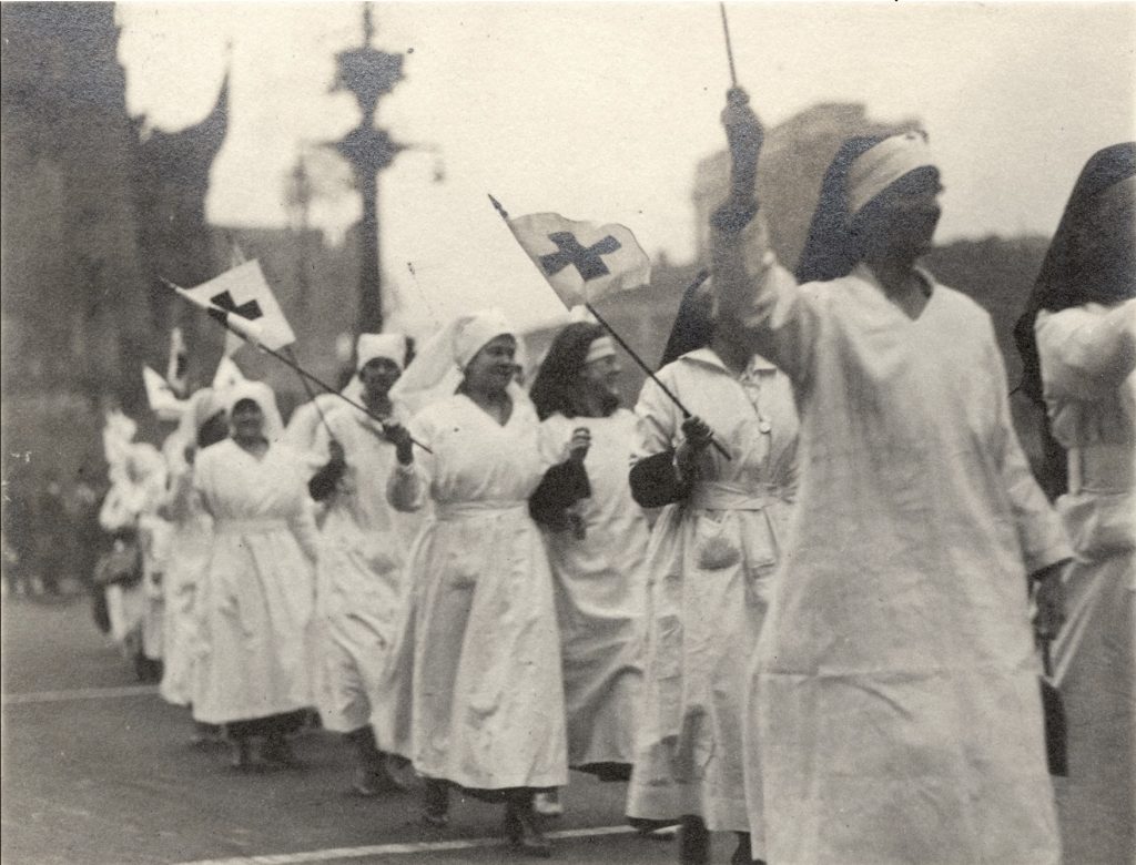 Women dressed in nursing uniforms and carrying flags with the red cross emblem parade around the circle. 