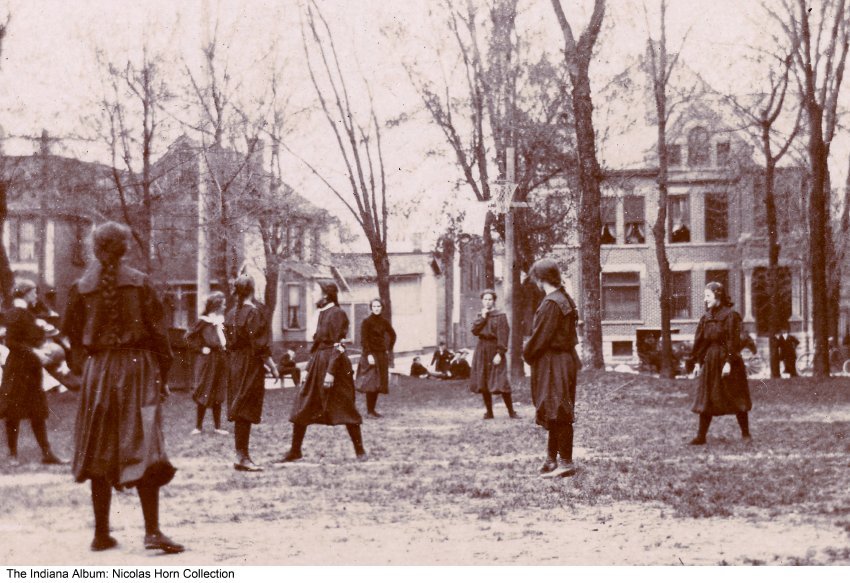 A group of girls in 1800s era basketball uniforms are spread across a field outside. 