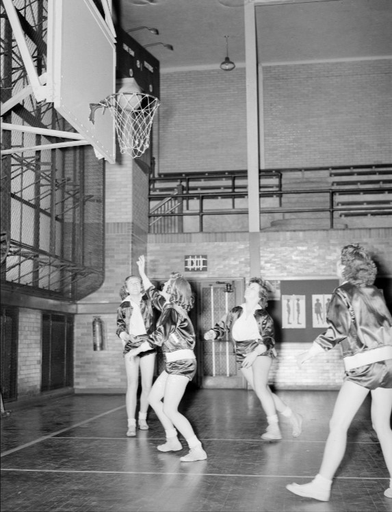 A player on the girls' basketball team at Shortridge tries to make a shot on the court. The team appears in their uniforms of satin jackets and shorts. This photo was taken by the 1939 Annual (yearbook) staff at Shortridge.