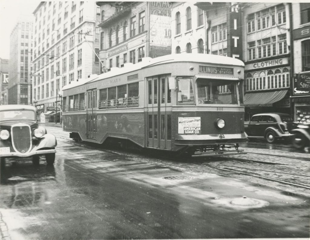 A trolley runs on a track in the middle of a city street. Buildings like the sides of the street.