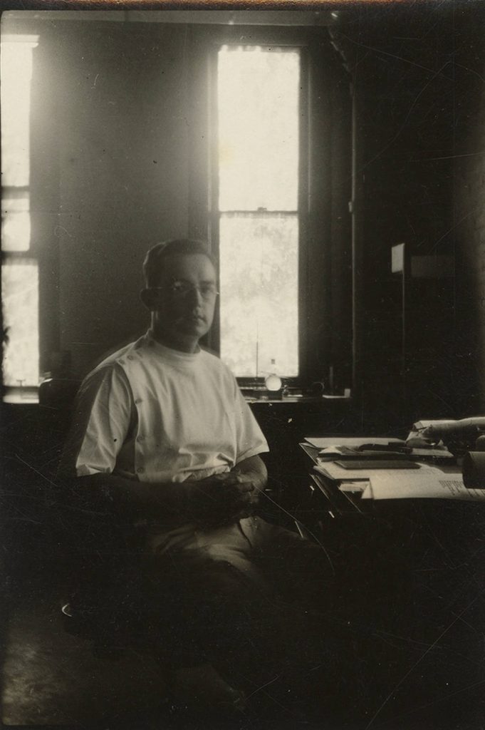 A man in a white uniform sits at a desk that is covered with papers.