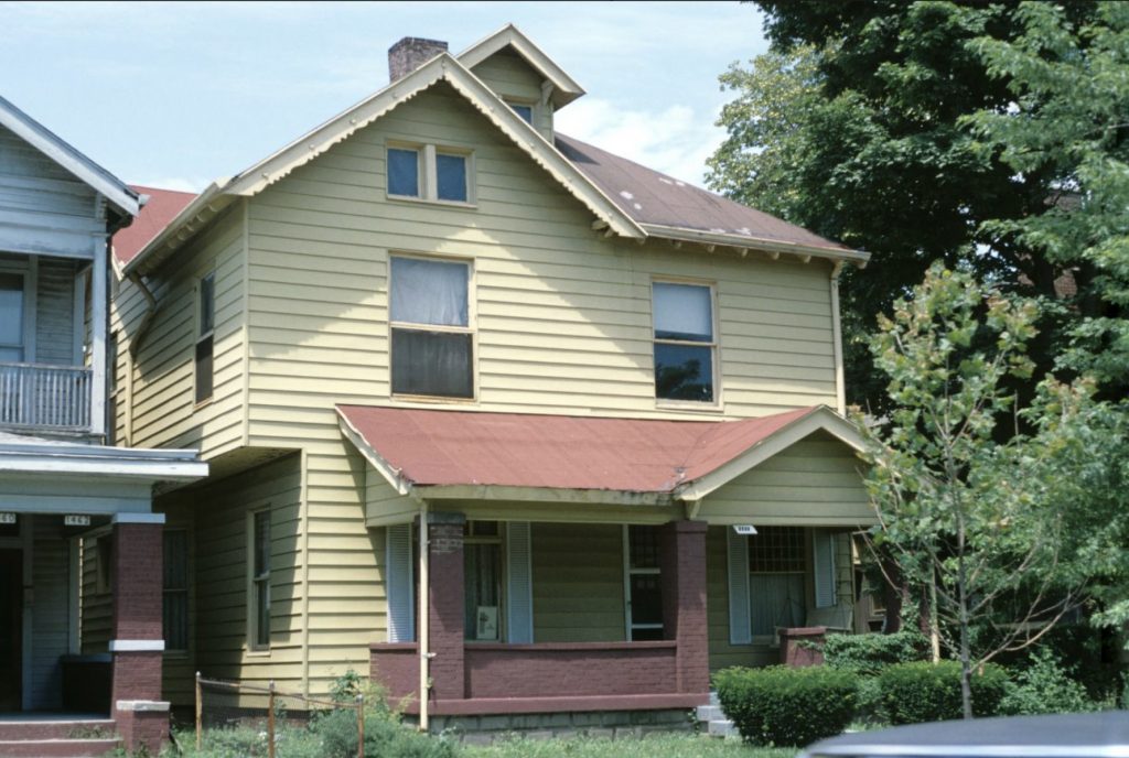 A multi-story Queen Anne-style house. A brick porch is attached to the front of the house. 