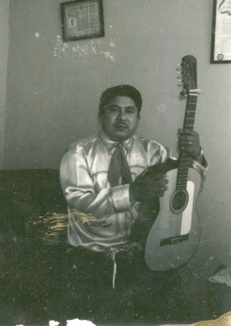 A man dressed in a silky shirt and neckerchief holds an acoustic guitar.