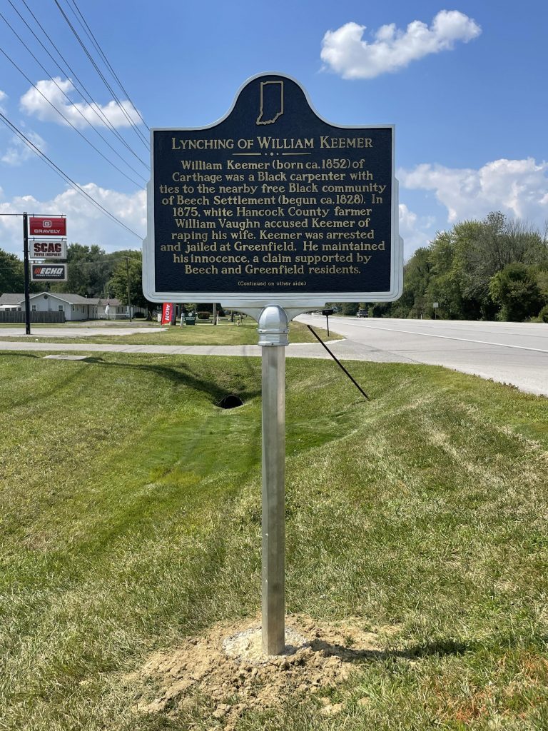 A historical marker sign stands in the middle of a patch of grass along a road. 