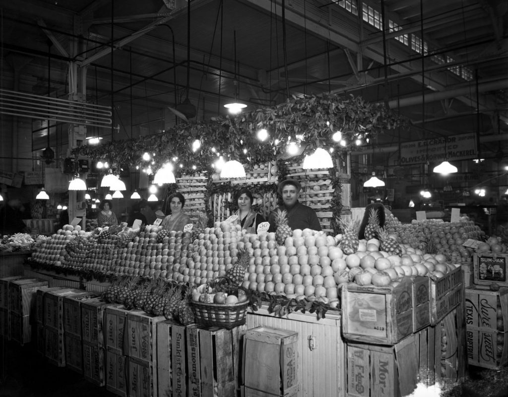 Three people stand behind the counter of a market stall. Fruit is stacked up in high piles in front of the people.