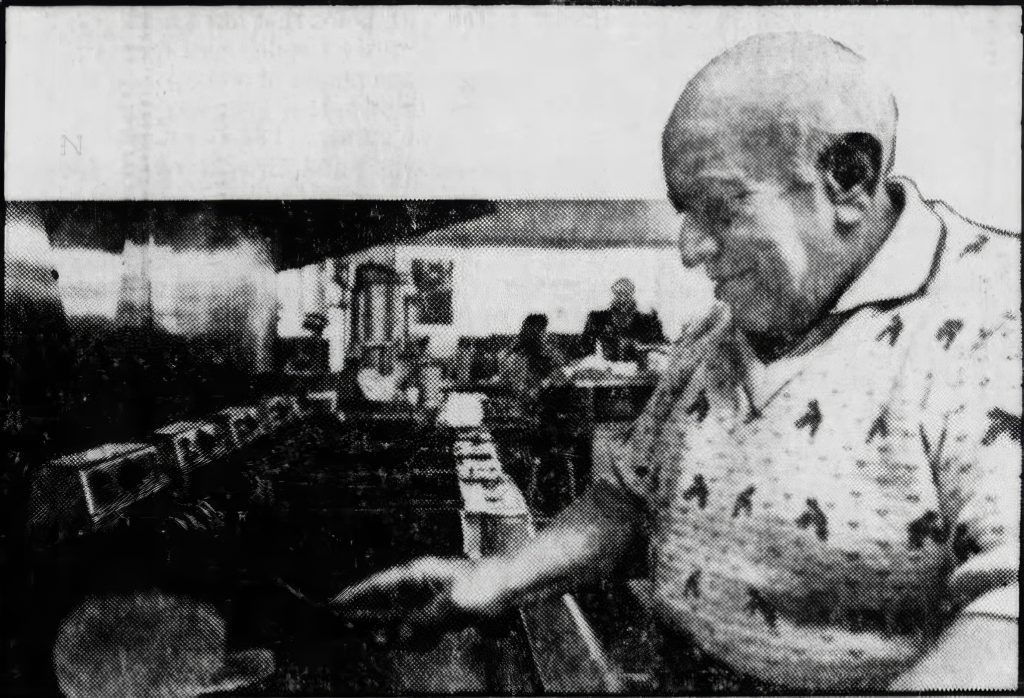 A man is flipping a hamburger on a grill. Customers are seated at a counter in the background.